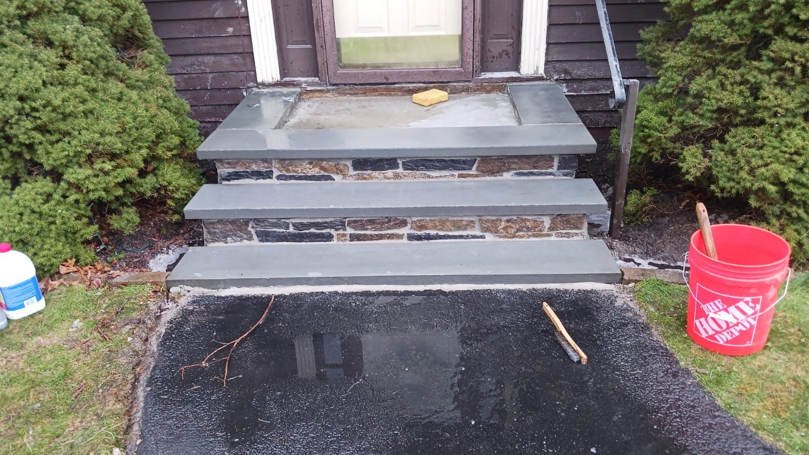 Stone steps leading to a house entrance. A bucket and brush sit nearby on a wet asphalt path.