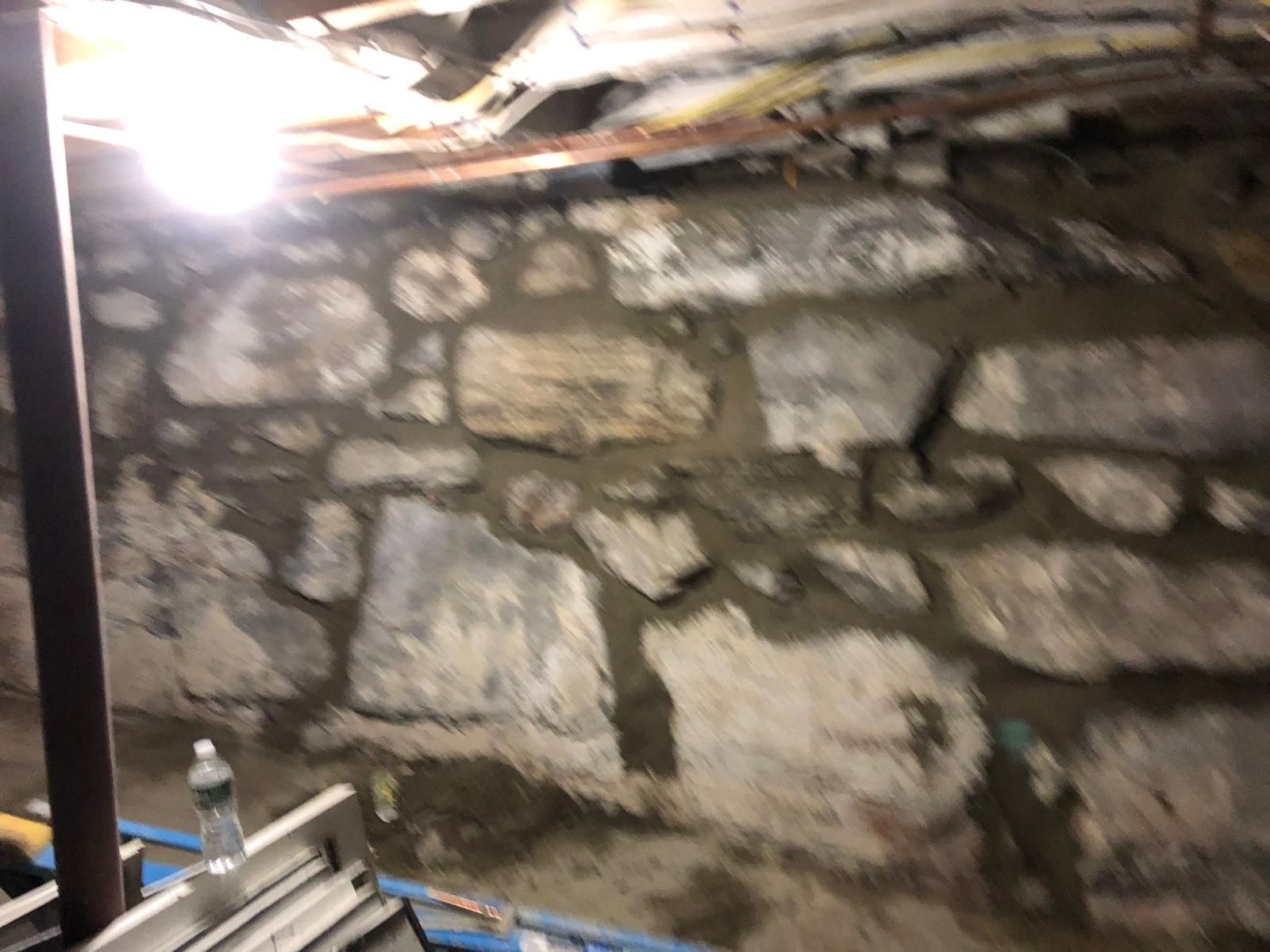 Stone foundation wall in a basement, with mortar, pipes and electrical wiring visible overhead.