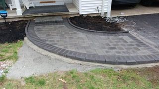 Brick paver pathway with dark border curving to porch. Black mulch and asphalt driveway in view.