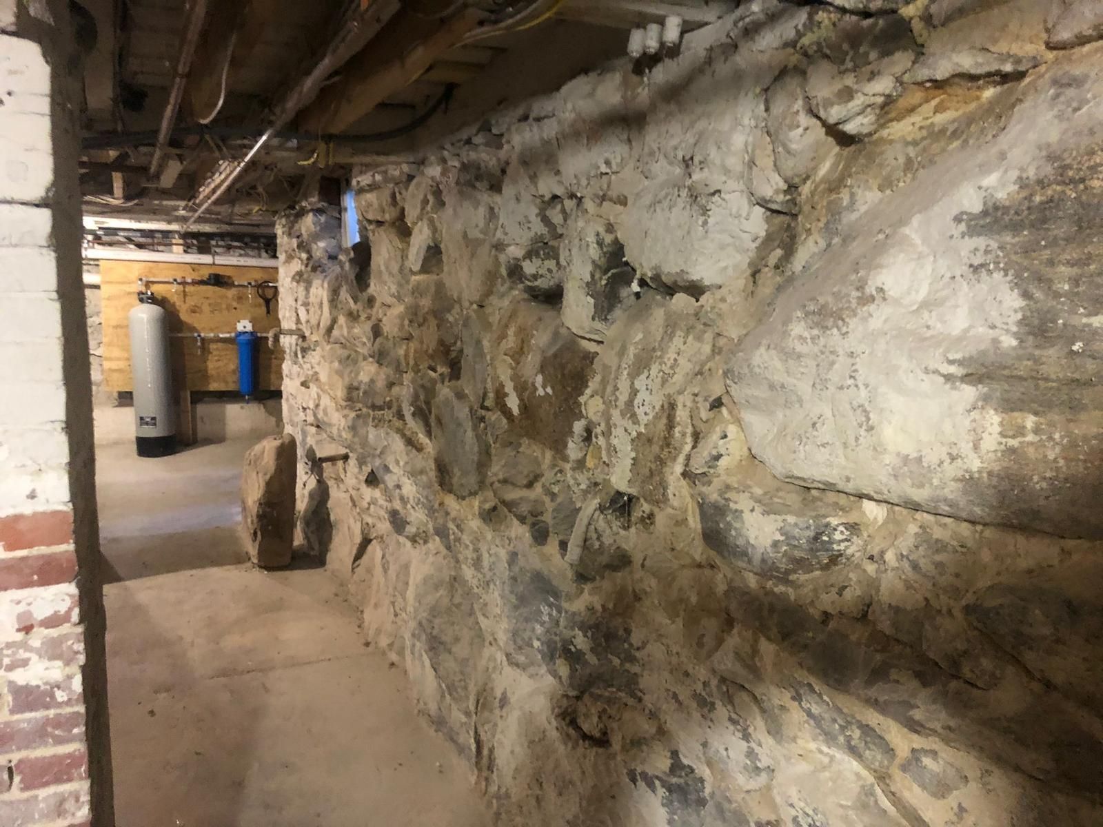 Stone foundation wall in a basement. Pipes and wires above, water softener and filters in the background.
