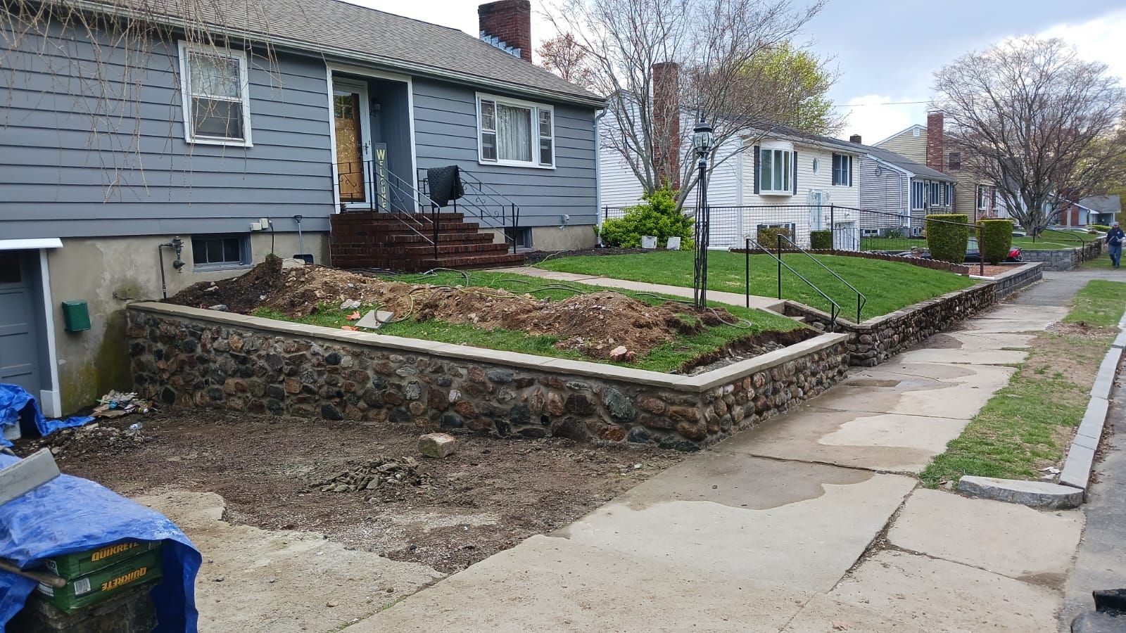 Gray house with stone retaining wall under construction; sidewalk in front.