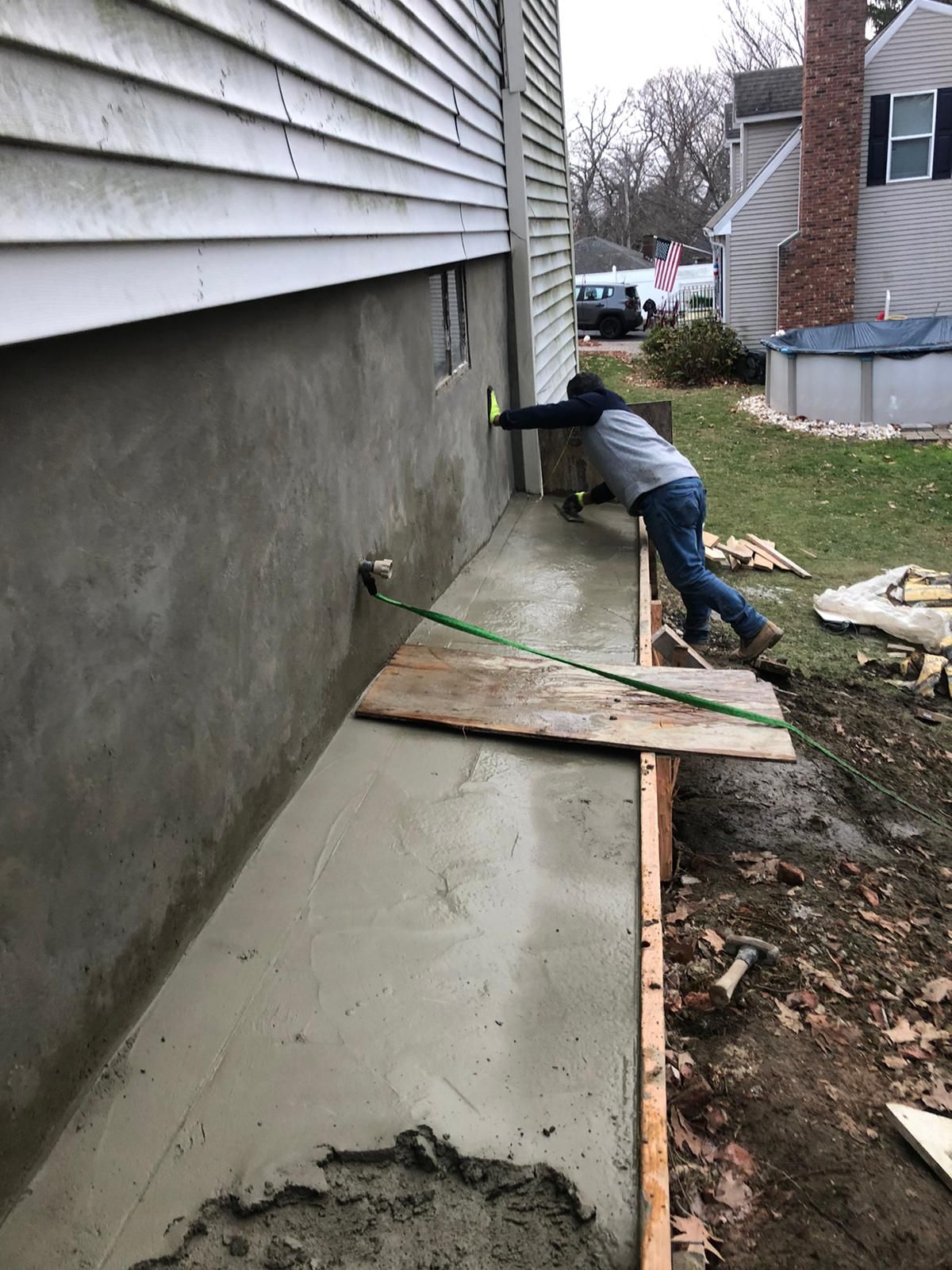 Man smoothing concrete on a walkway next to a house. Gray siding, wooden forms, and a green strap are visible.