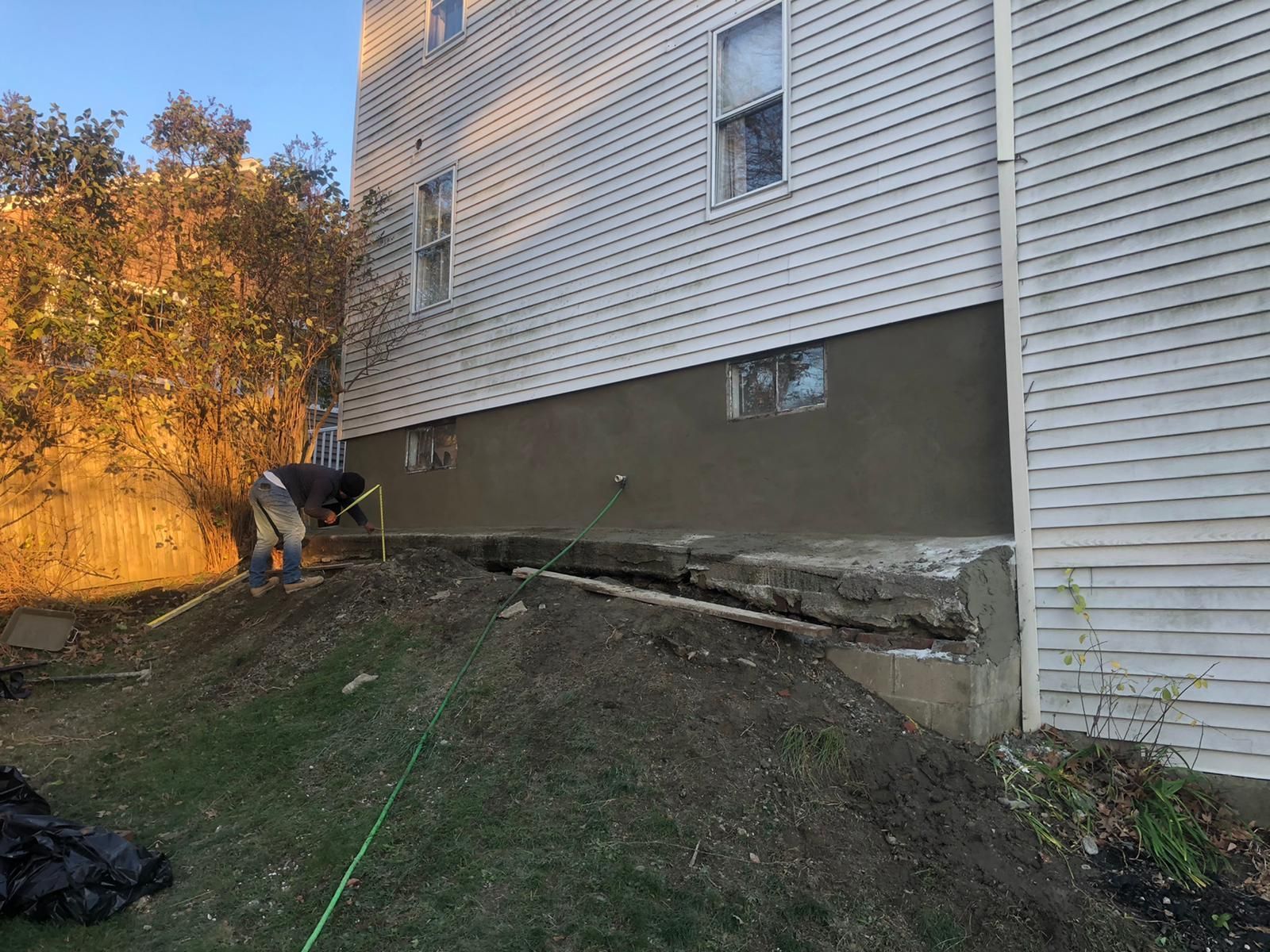 A person works on a building's foundation. The house is white with a gray base, in an outdoor setting.