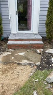 Brick steps leading to a white doorway. Damaged concrete path in front, dirt and grass around.