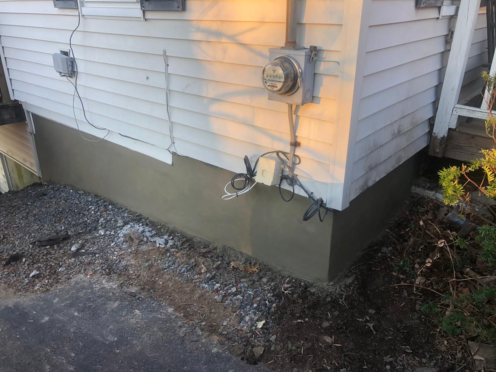 Tan stucco foundation on a white house, with electrical meter and wiring, next to gravel.