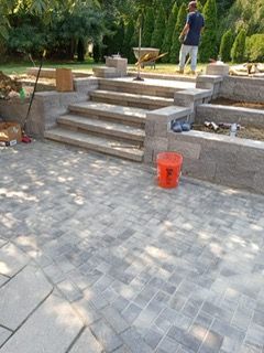 Patio with steps, gray pavers, and a worker in the background. Orange bucket in the foreground.