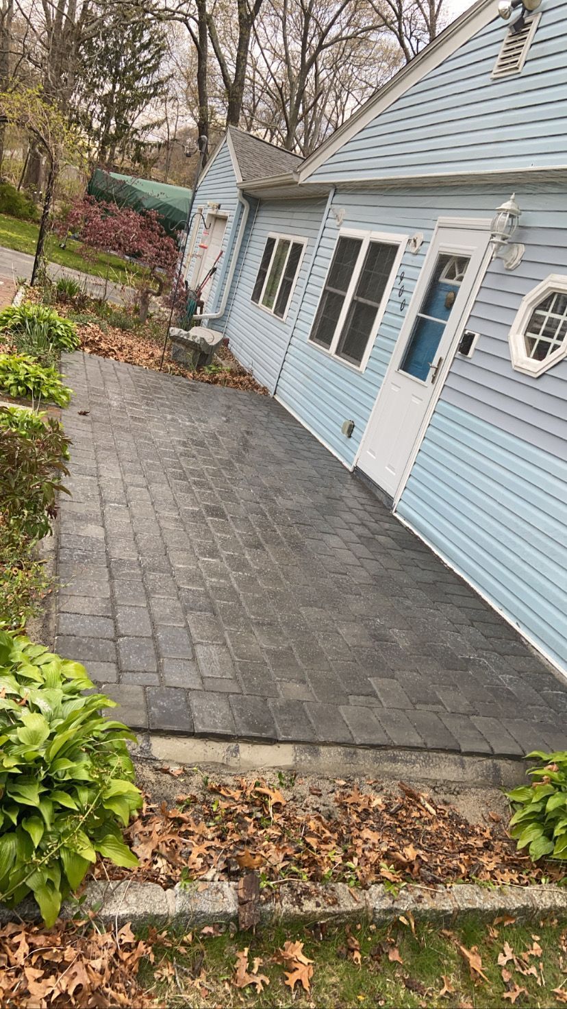 A brick patio next to a light blue house with windows, a door, and green bushes.