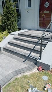 Gray brick steps and walkway lead to a house entrance with a black handrail.