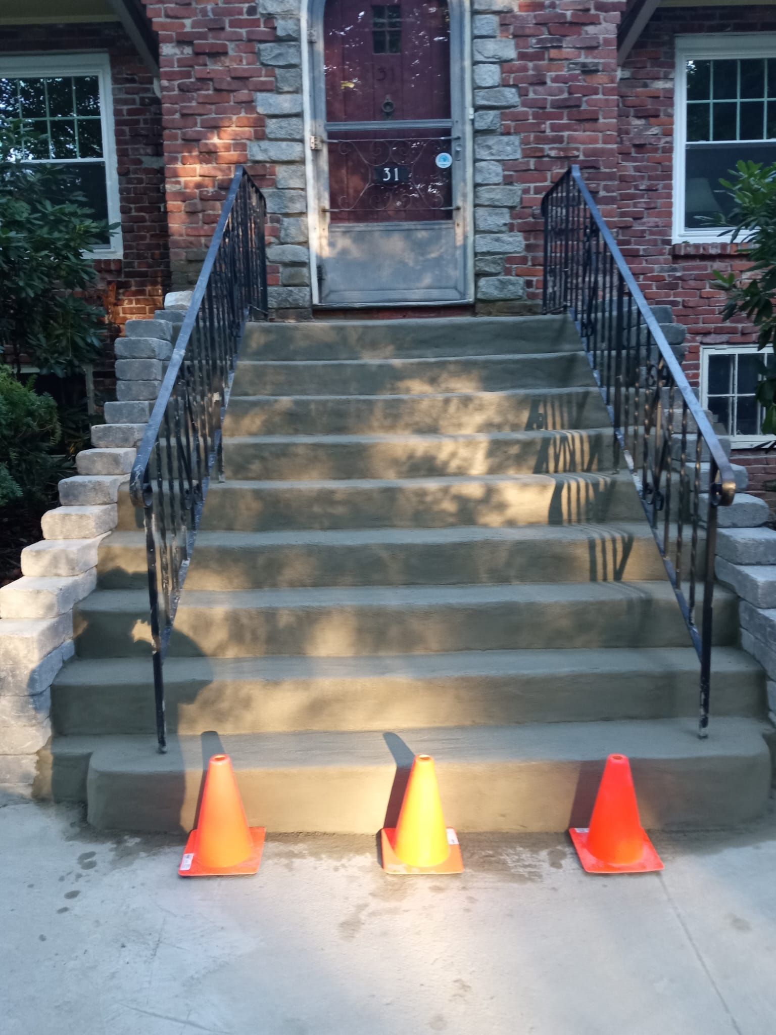 Concrete steps leading to a brick house, with black railings and three orange traffic cones at the bottom.