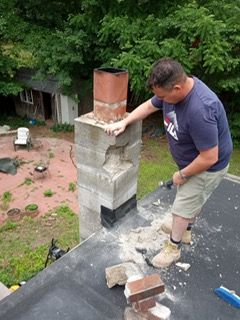 Man on a roof, chiseling at a damaged concrete chimney. Outdoors, trees and a building in the background.