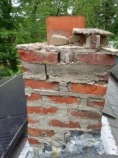 A weathered brick chimney with crumbling mortar, atop a rooftop with a rusted metal base.