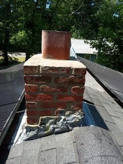 Brick chimney on a dark shingle roof with deteriorating flashing; a red metal flue is on top.