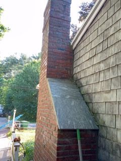 Brick chimney next to a shingled house; angled siding section; green trees in the background.