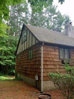 Brown shingled house with a steeply pitched roof in a wooded area, with a chimney and a small window.