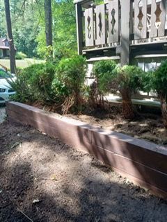 Brown raised garden bed with newly planted green shrubs in front of a wooden fence, outdoors.