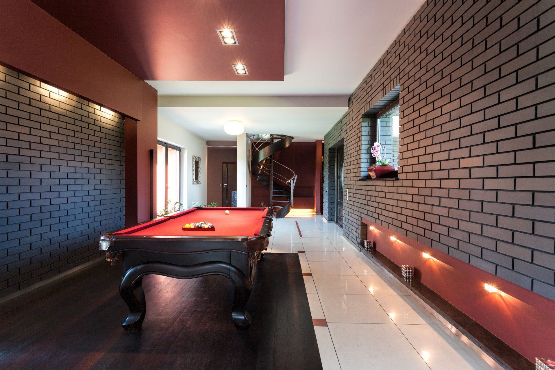 Pool table in a long, modern room with brick accent wall, burgundy accents, and spiral staircase.