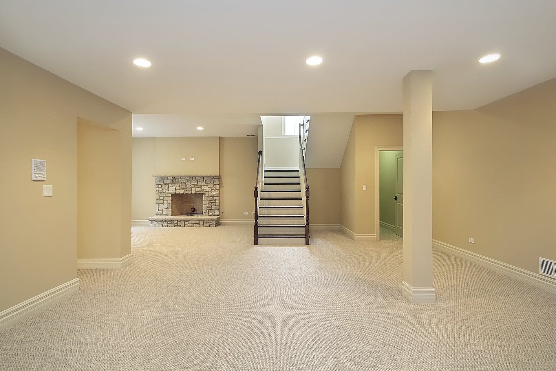 Empty basement with carpet, beige walls, fireplace, and stairs.