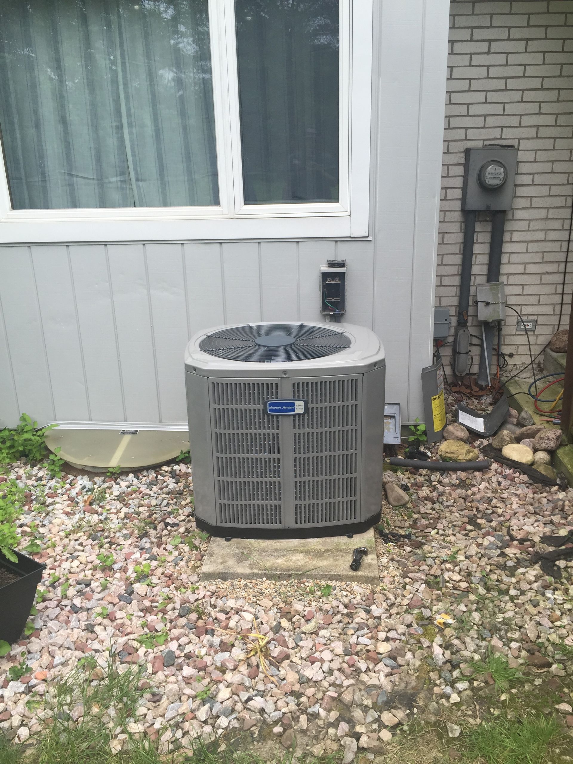 Outdoor air conditioner unit beside a house window, surrounded by gravel and leaves.