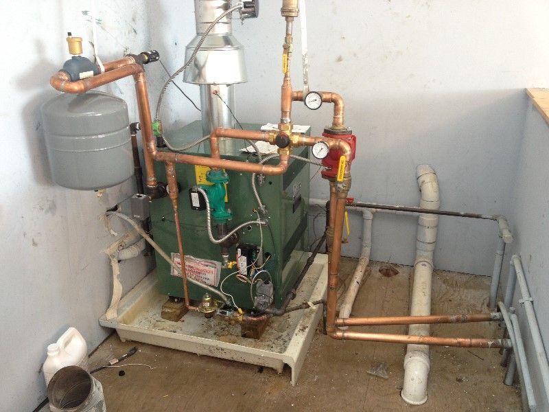 Basement utility room with green boiler, copper pipes, and gray expansion tank beside concrete floor.