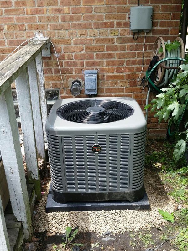 Outdoor air conditioner unit beside a brick wall and wooden railing in a small yard