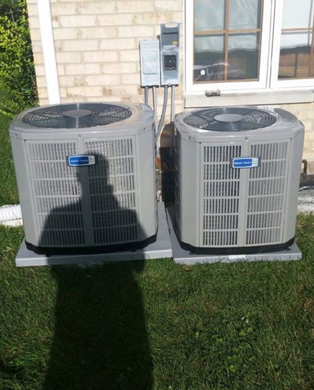 Two gray outdoor air conditioner units beside a house, with a person’s shadow on the grass.