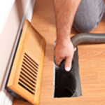 Hand holding a black air duct filter beside a wooden floor vent cover