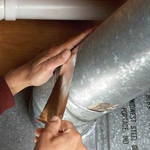 Hands taping a seam on silver ductwork with brown tape and a tool.