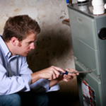 Technician inspecting a furnace with a tool, kneeling beside a gray heating unit.