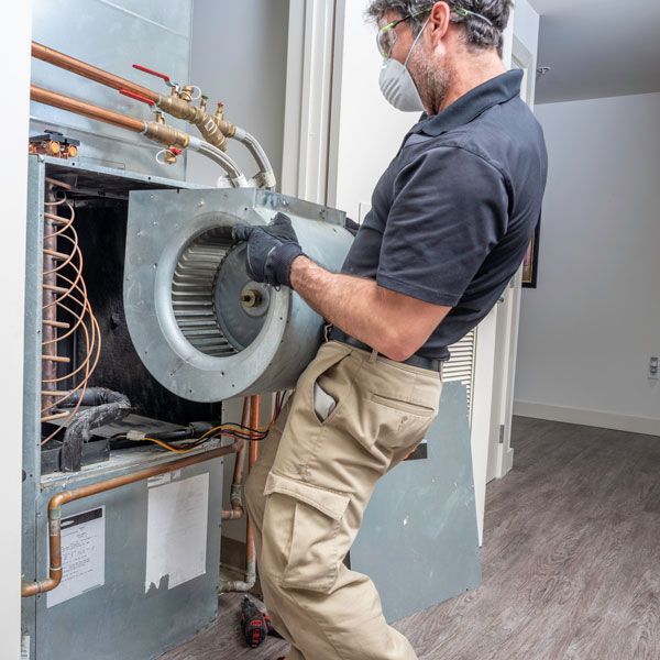 Technician wearing a mask and gloves servicing an HVAC unit indoors with exposed wiring and metal panels