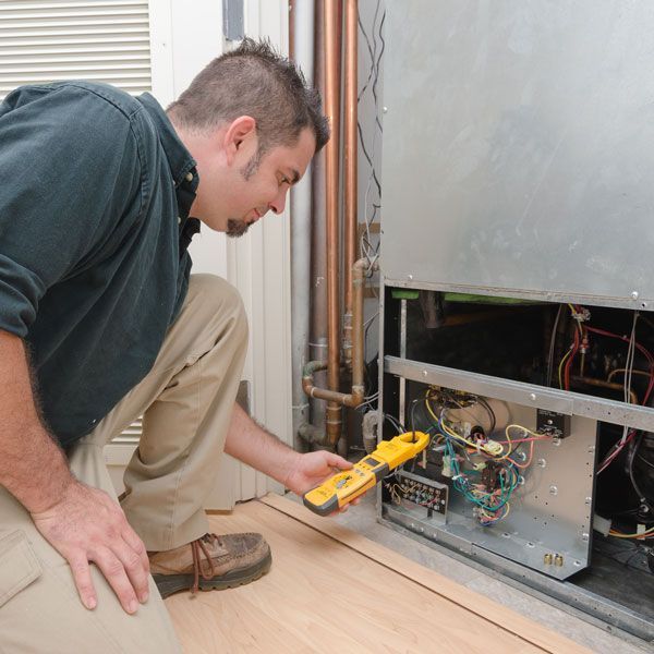 Technician kneeling by an open HVAC unit, using a yellow multimeter to check wiring.