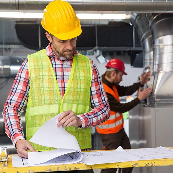 Construction workers reviewing blueprints in an industrial building, one in a yellow hard hat in front and one in red behind.