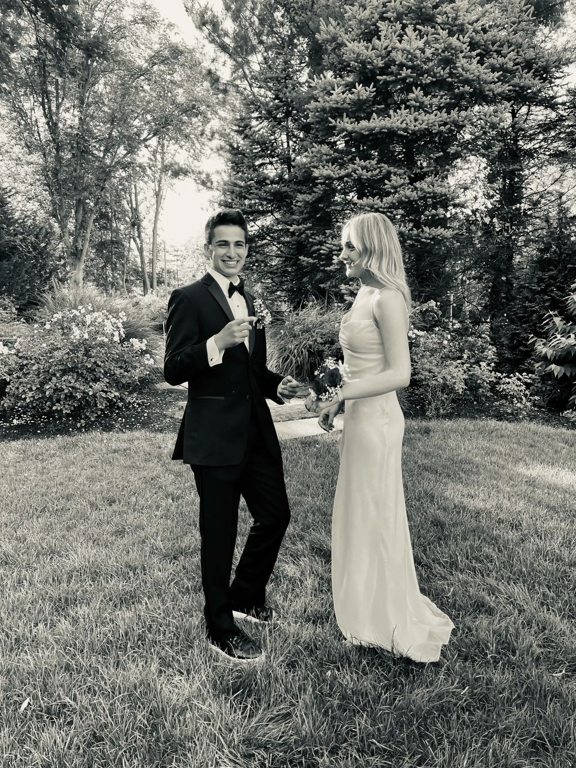 A couple in formal attire stands in a garden. The man holds a boutonniere, smiling, as the woman looks on.