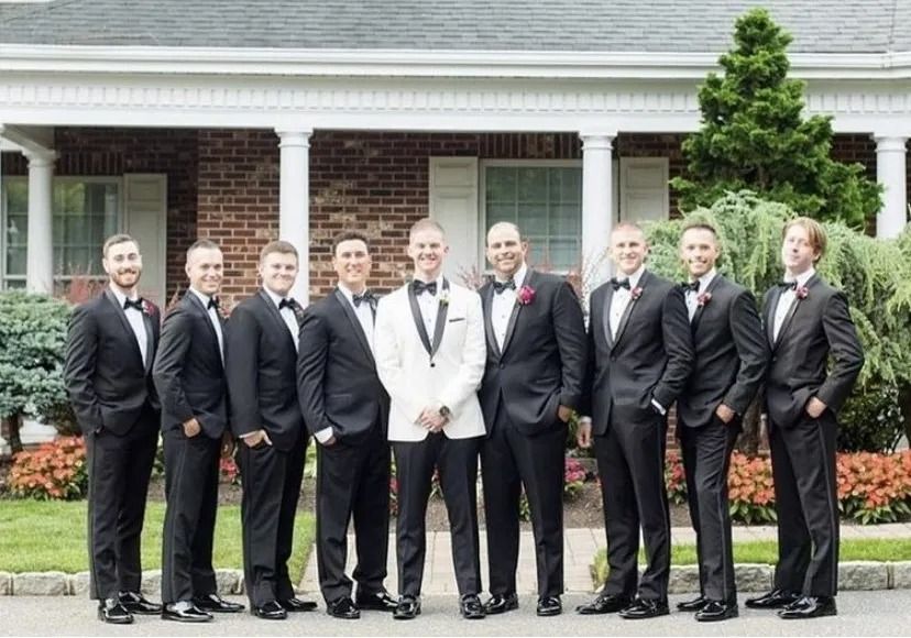 Nine men in tuxedos pose in front of a house; one wears a white jacket.