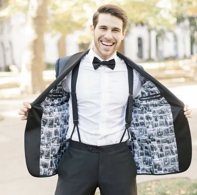 Man in tuxedo with suspenders, smiling, opening jacket to reveal printed lining. Outdoor setting.