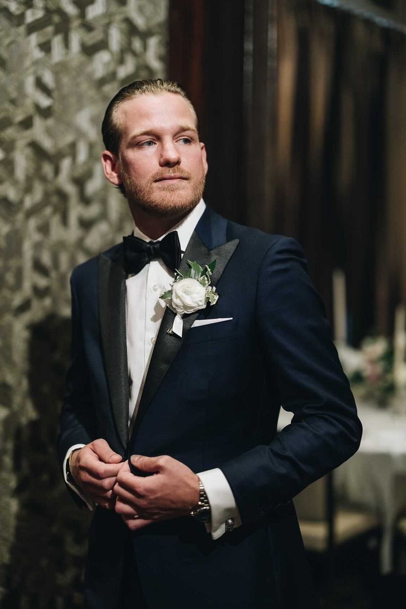 Man in navy tuxedo, black bow tie, white shirt, and boutonniere; indoor setting, looking away, buttoning jacket.