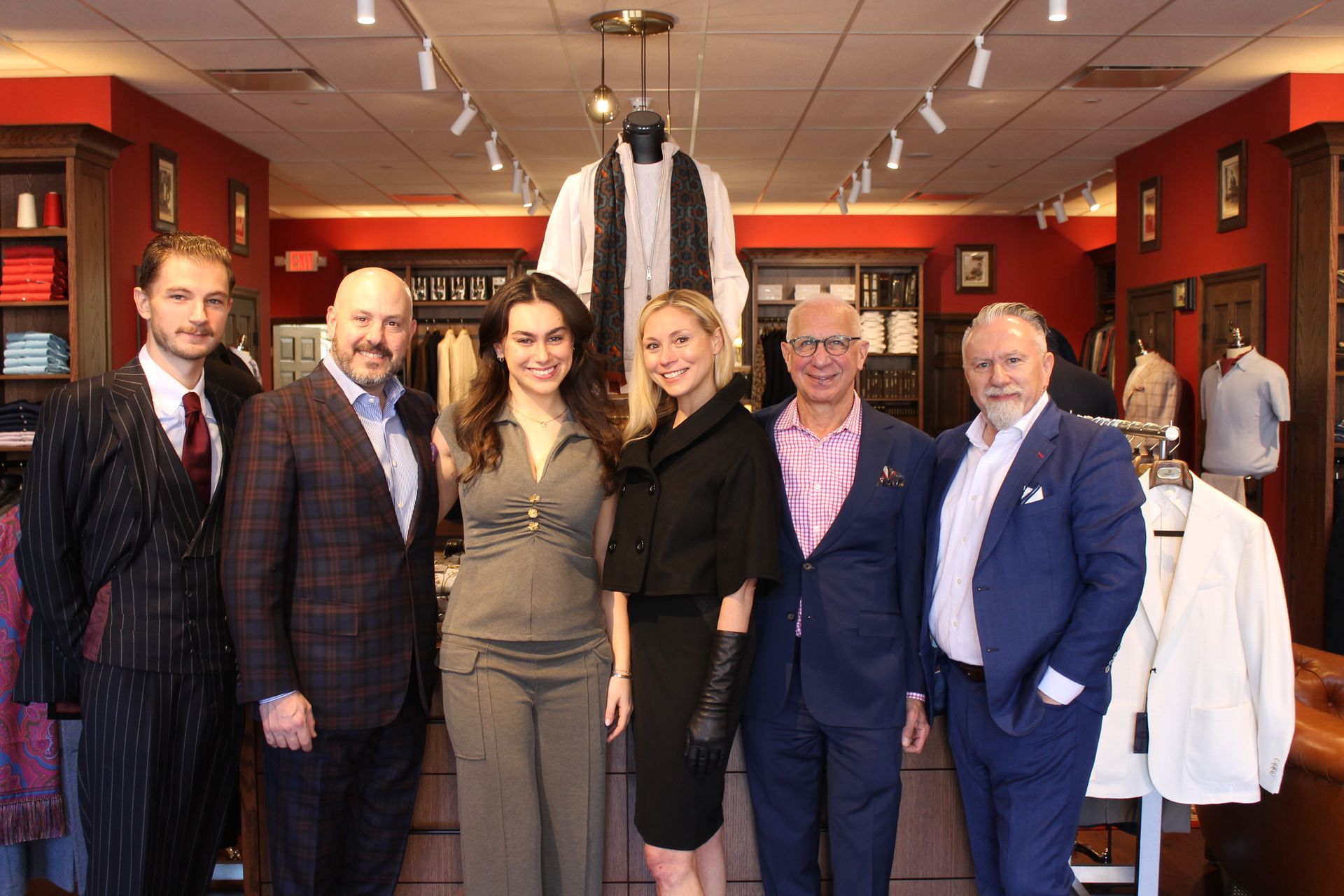 A group of six people stand smiling inside a well-lit clothing store, posing for a photo in front of racks of apparel.