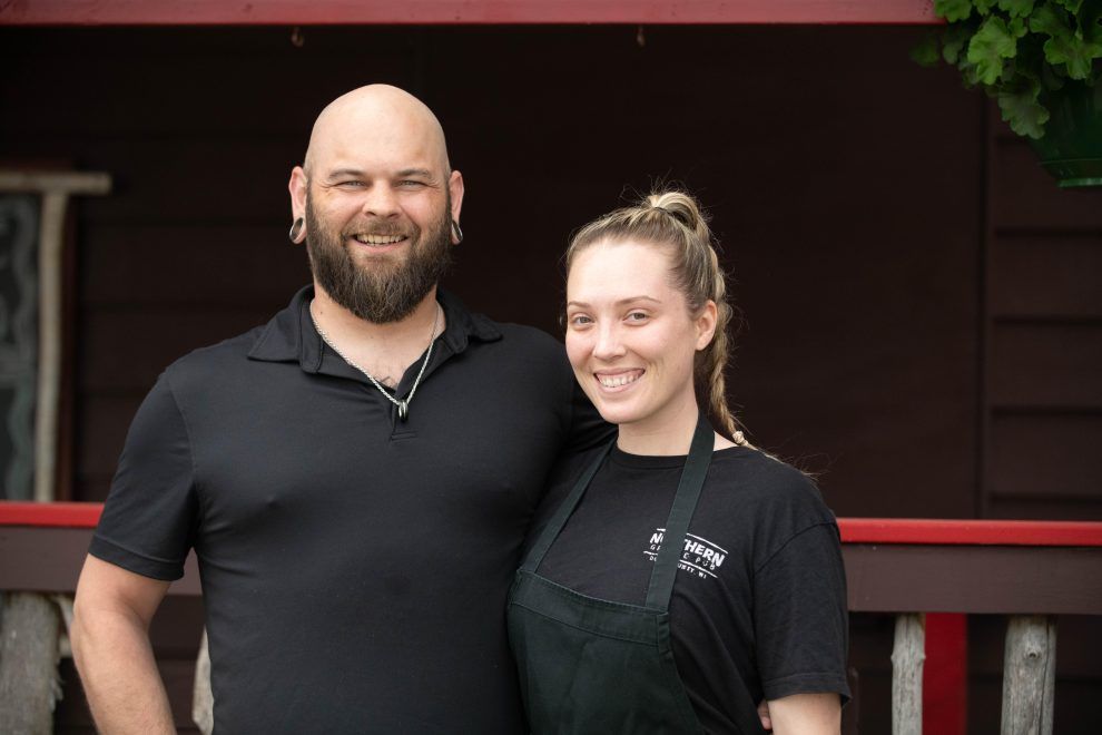 Two people smiling and standing together outdoors in front of a dark wooden building.
