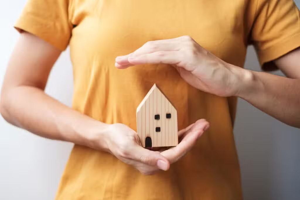 Hands holding a small wooden house model against a yellow shirt background
