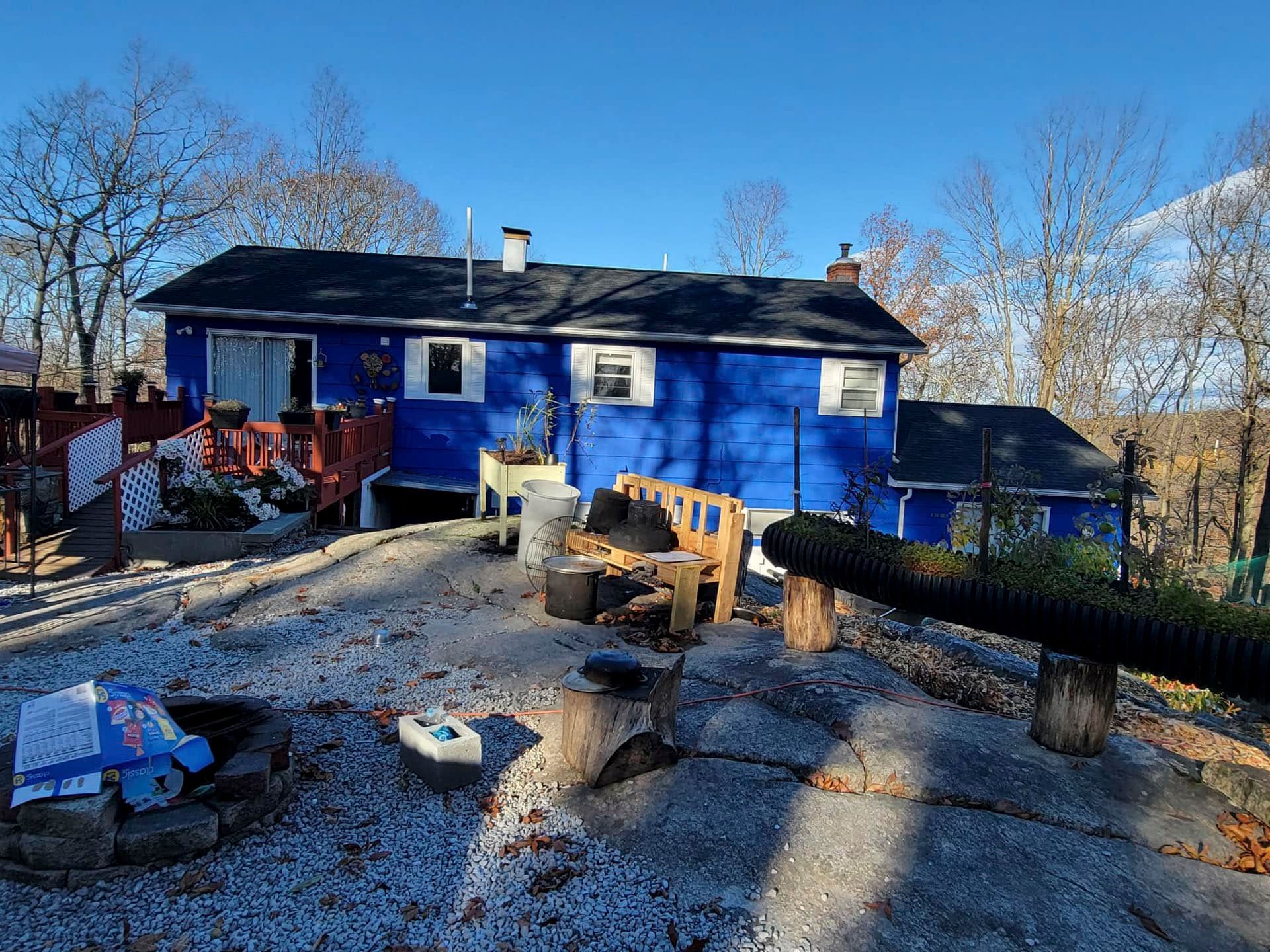 Blue house with a front deck, fire pit, and chairs in a sunny yard with bare trees.