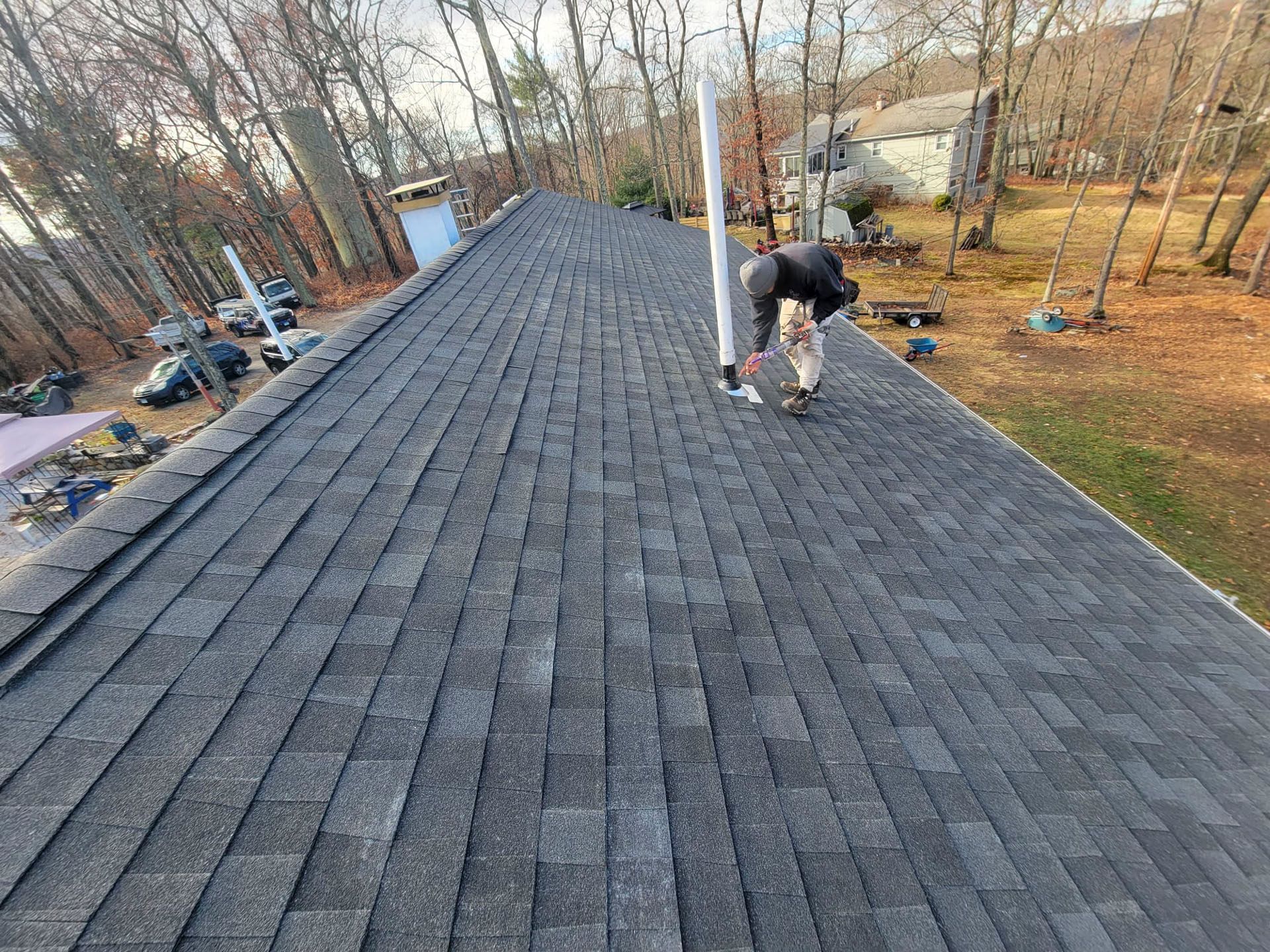 Worker installing shingles on a steep roof beside trees and a house