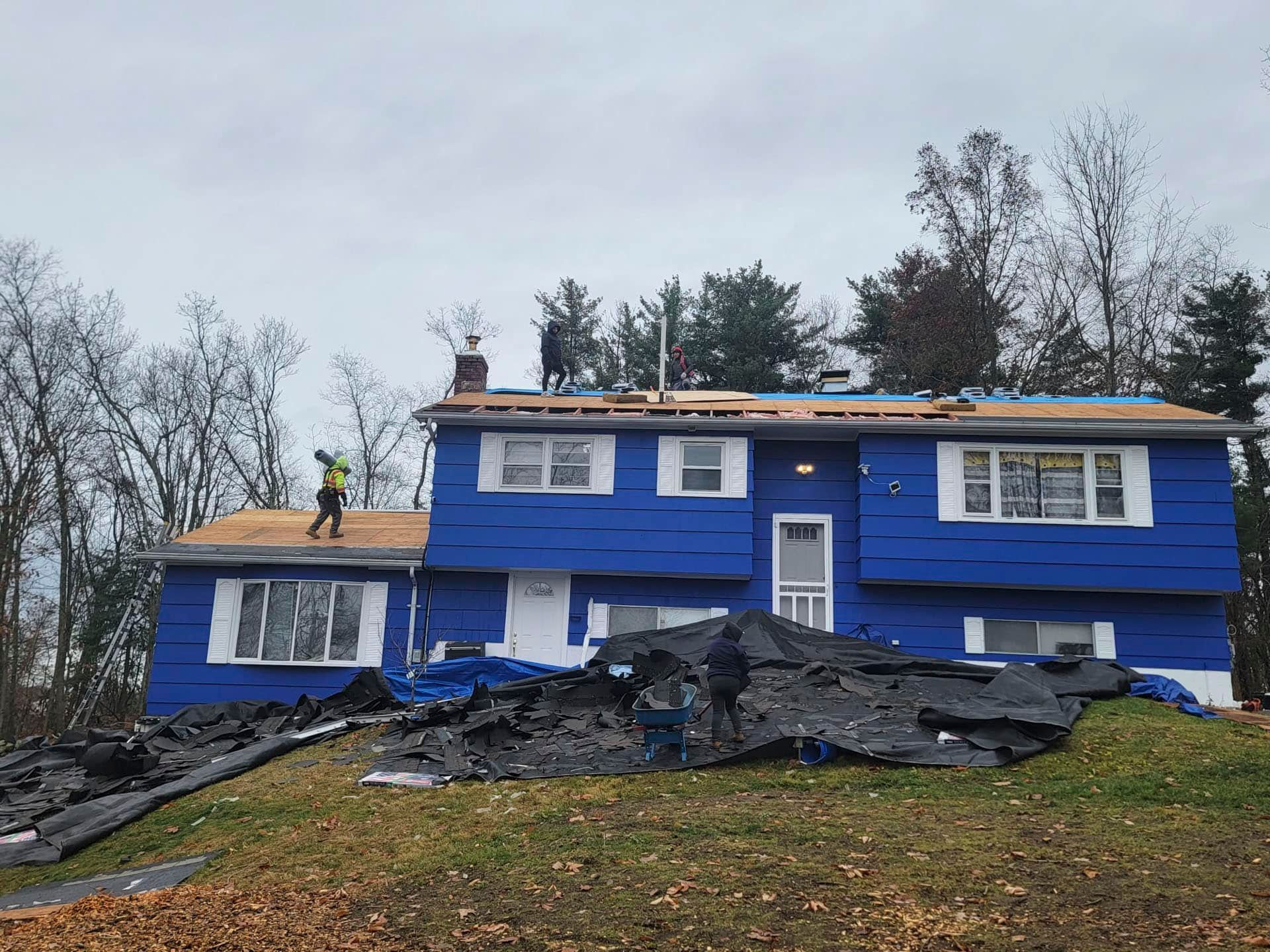 Blue house with roof damage and debris piled in front, set on a grassy slope with leafless trees behind.
