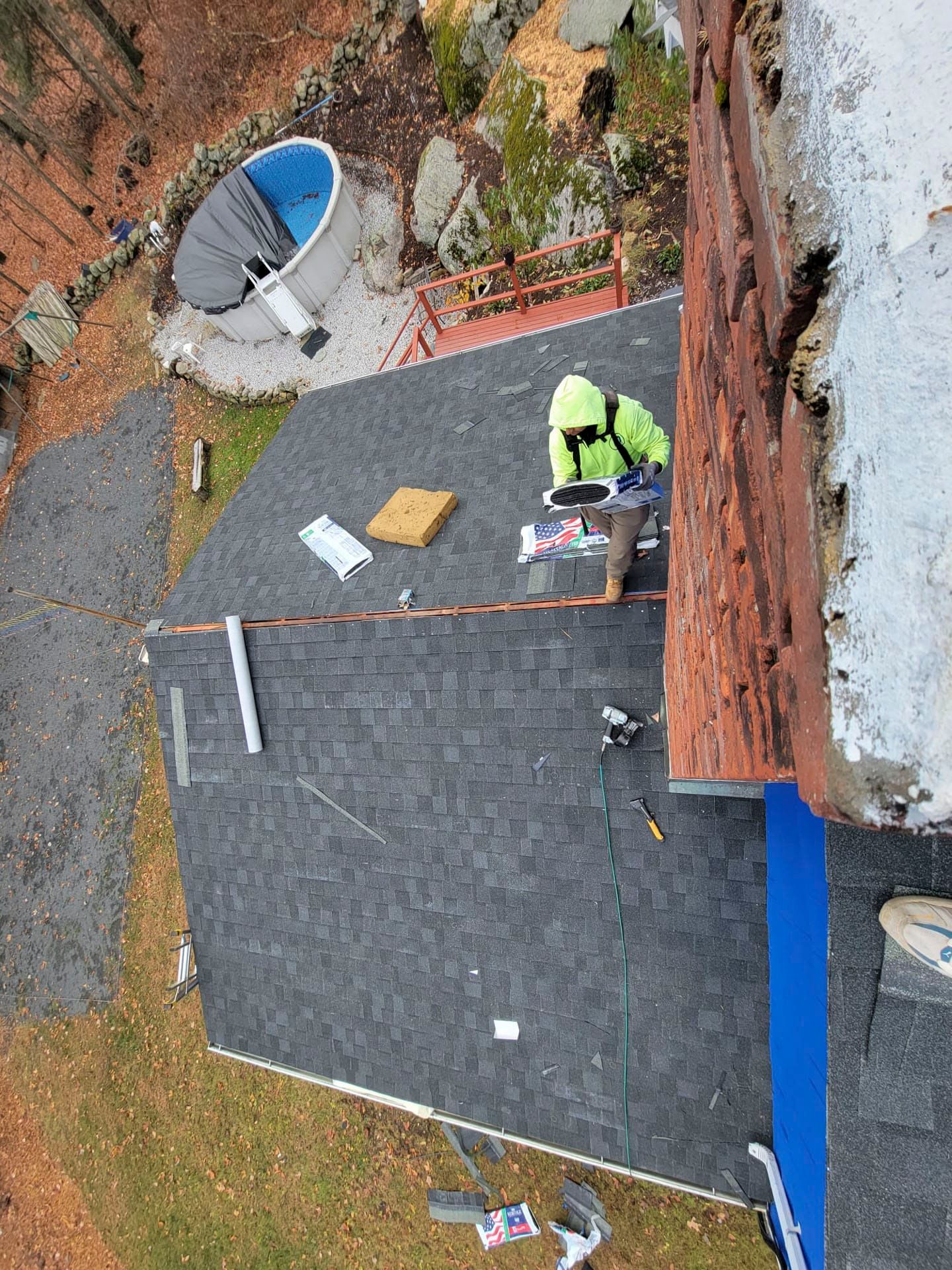 Worker in neon jacket on a rooftop, with tools and materials beside a sloped gray roof.
