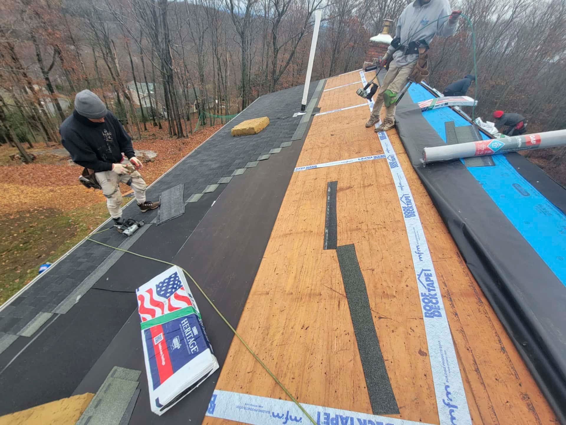 Workers installing roofing on a sloped roof, with tar paper, blue underlayment, and trees in the background