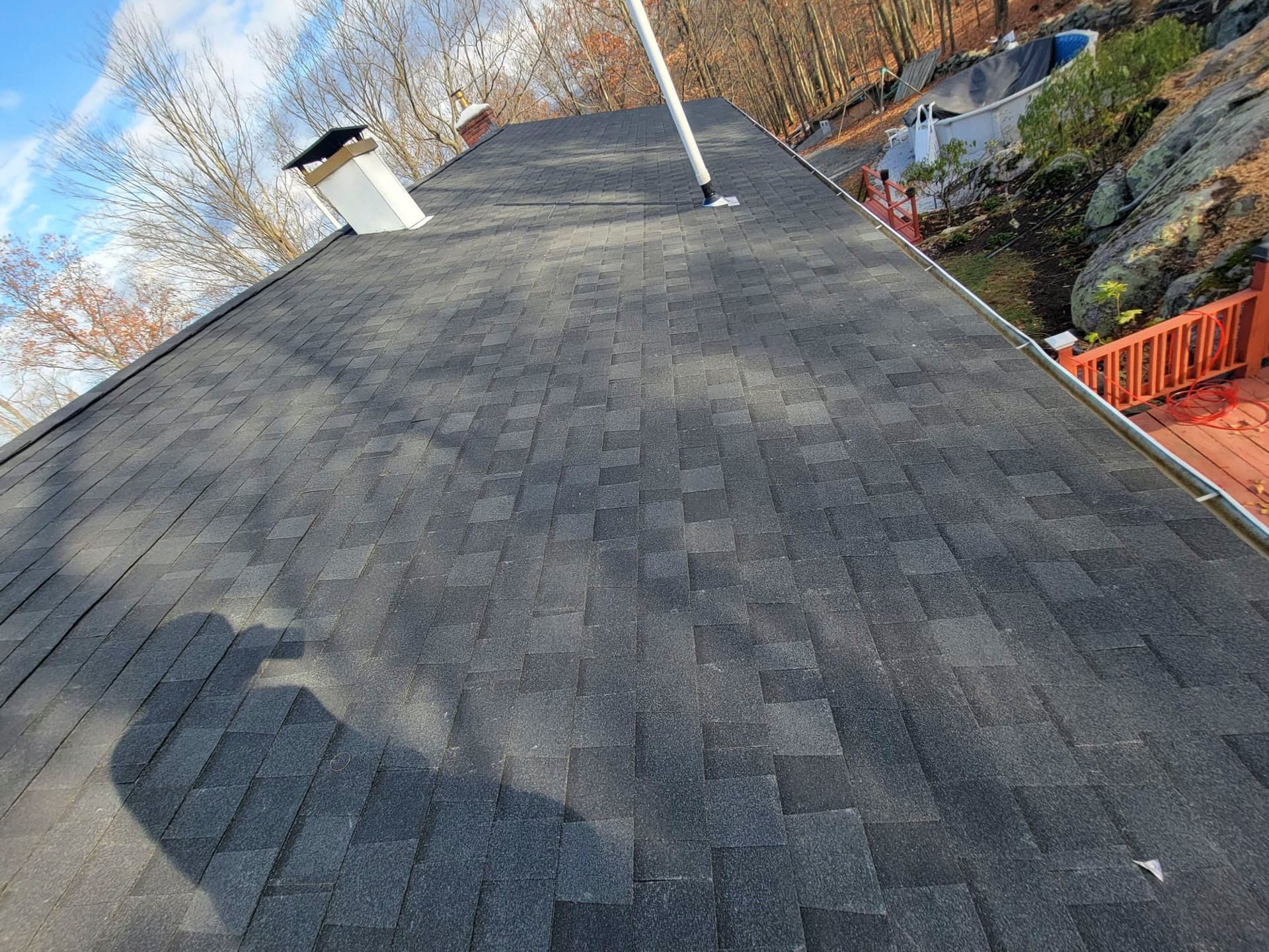 Dark gray shingled roof with a chimney and rooftop vent, viewed at an angle beside trees and a street below