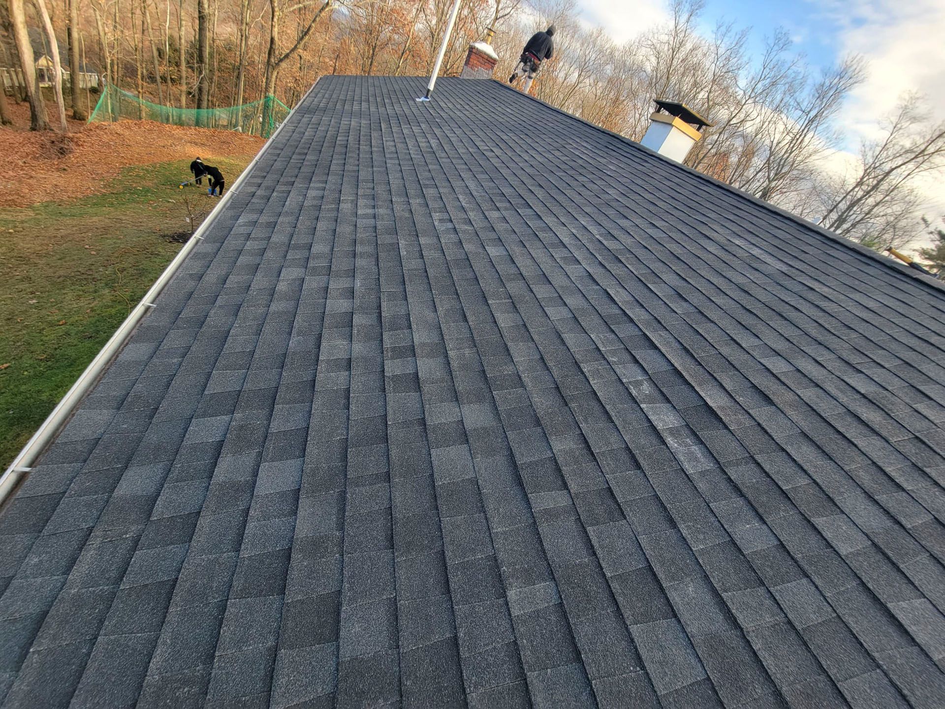 Close-up of a gray shingled roof on a house, viewed at an angle with trees in the background