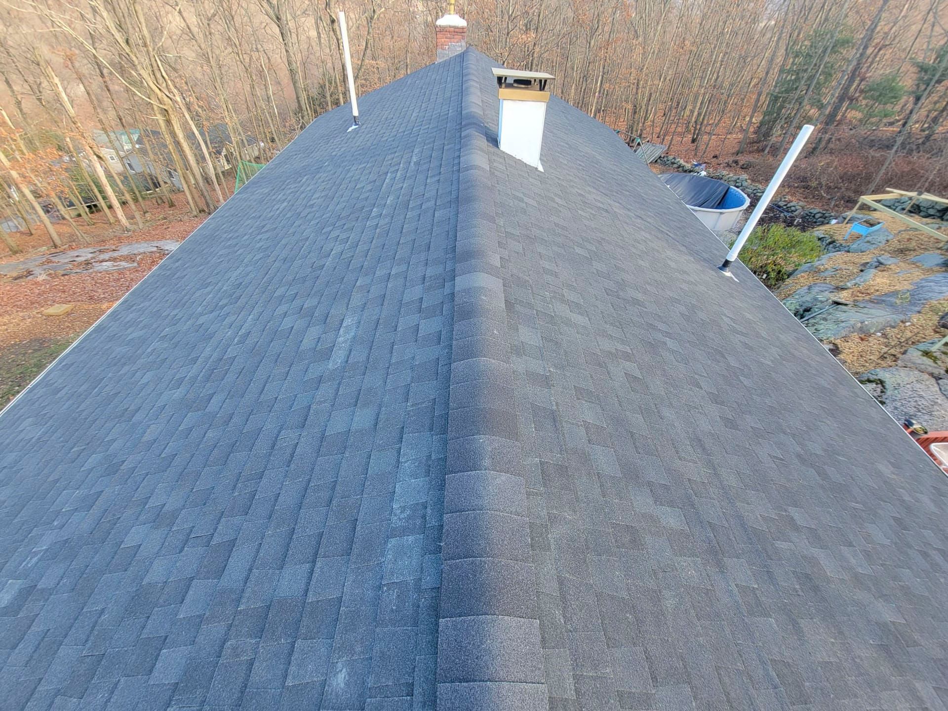 Aerial view of a dark shingled roof with a central ridge, chimney, and vent pipes near wooded ground.