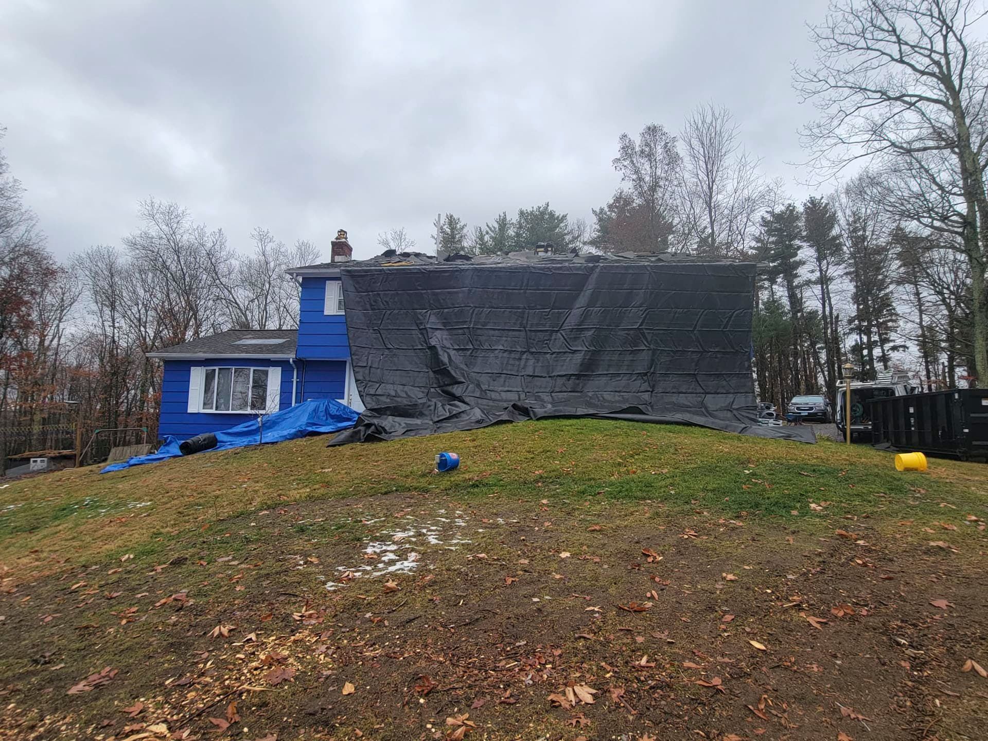 Blue house with tarp-covered roof on a grassy hill under cloudy skies