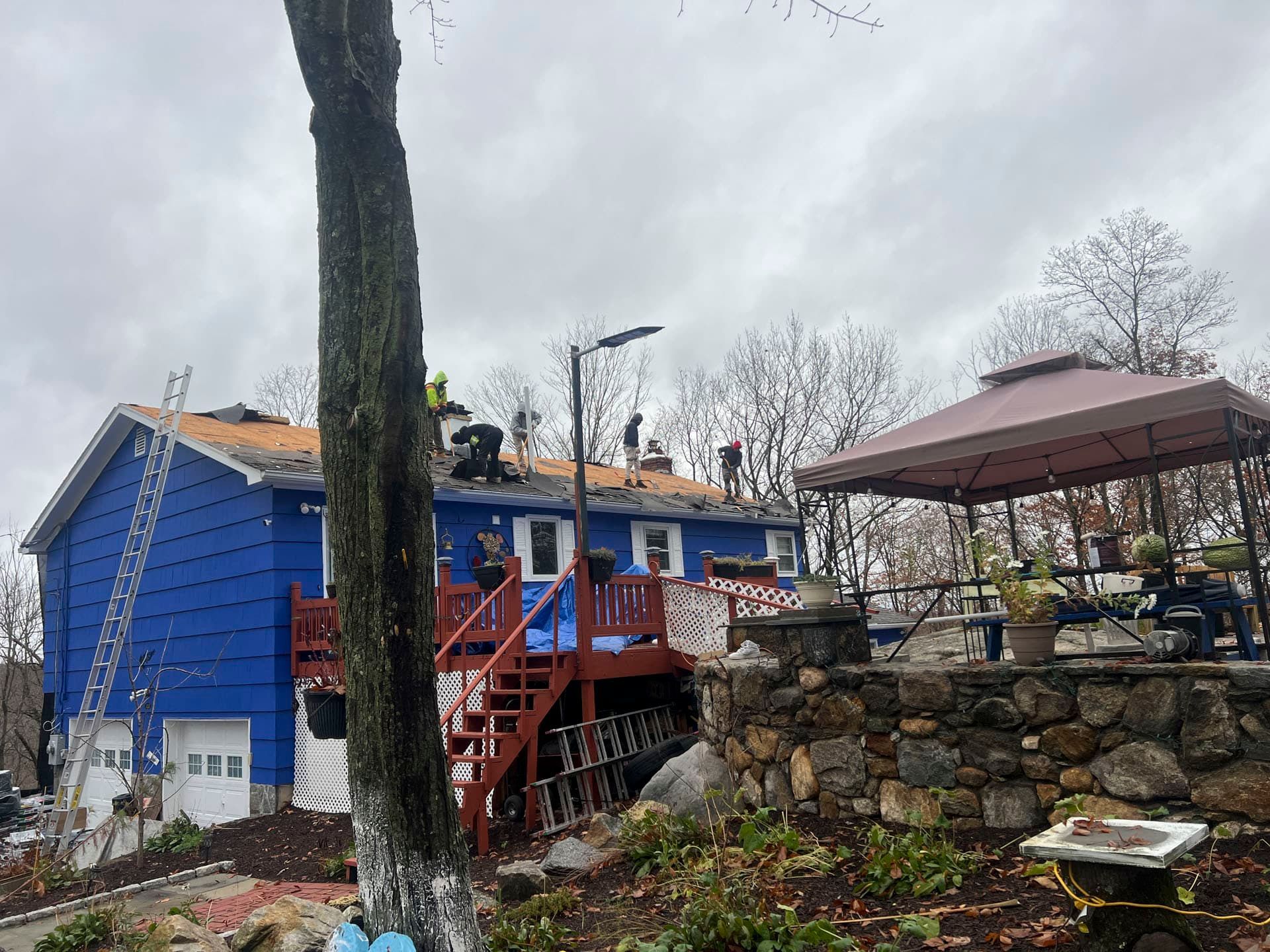 Blue house with red stairs on a stone hillside, with a rooftop patio and gazebo under a cloudy sky