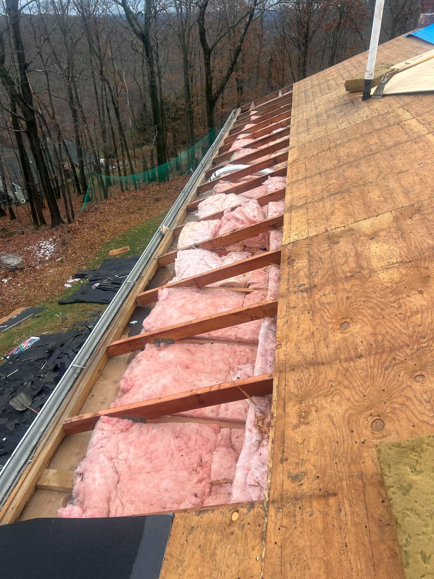 Roof under construction with exposed pink insulation and plywood decking along the eaves on a wooded slope.