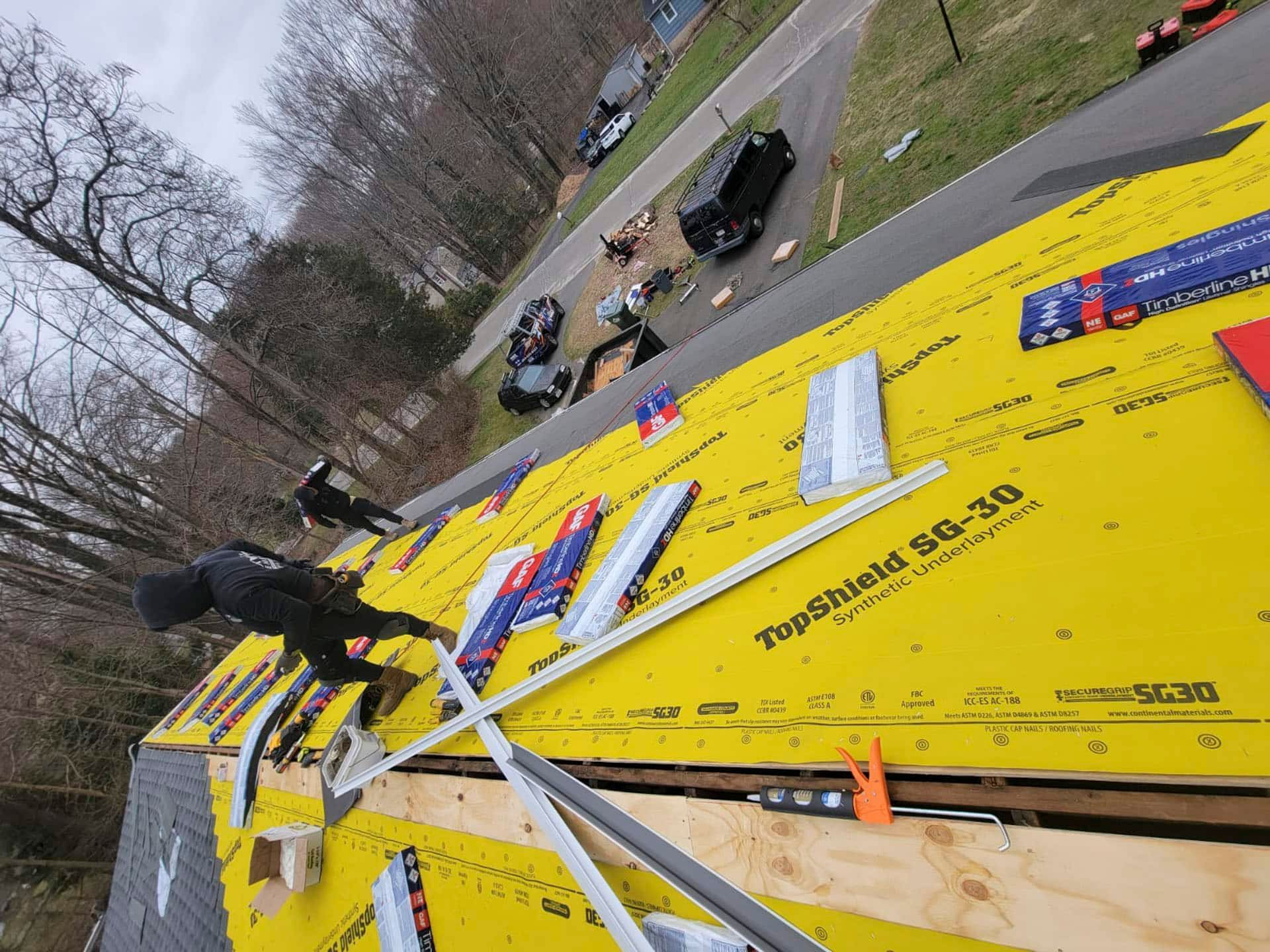 Roofing materials and tools on a steep yellow roof, with workers below near a road and trees